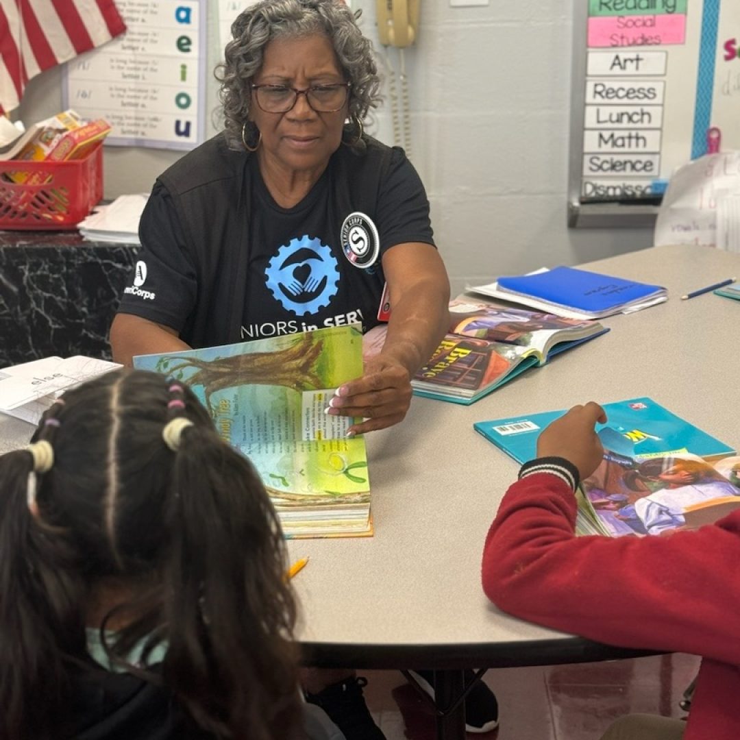 Seniors in Service Classroom Grandparent volunteer tutoring students on reading comprehension with book.