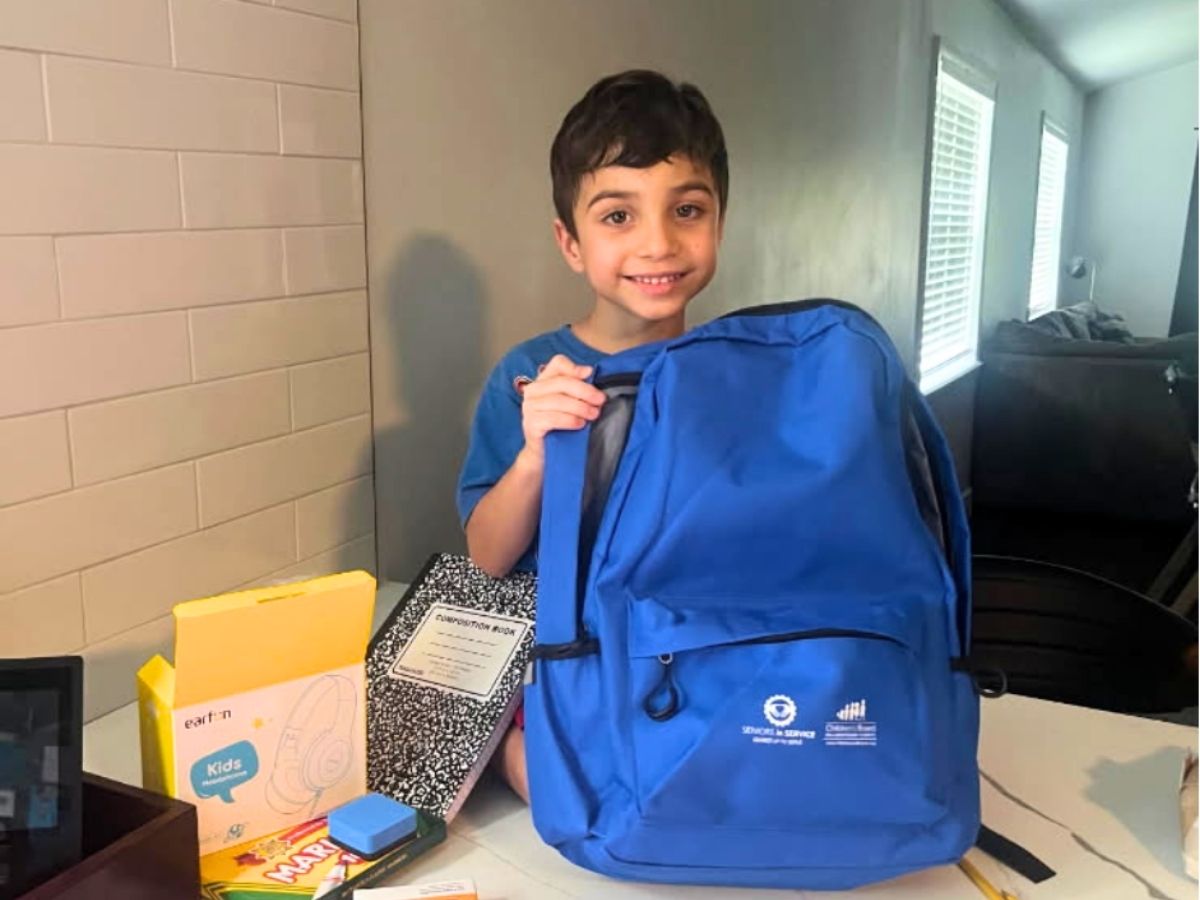 Young student smiling while holding a new backpack filled with school supplies provided through the Classroom Grandparent Program.