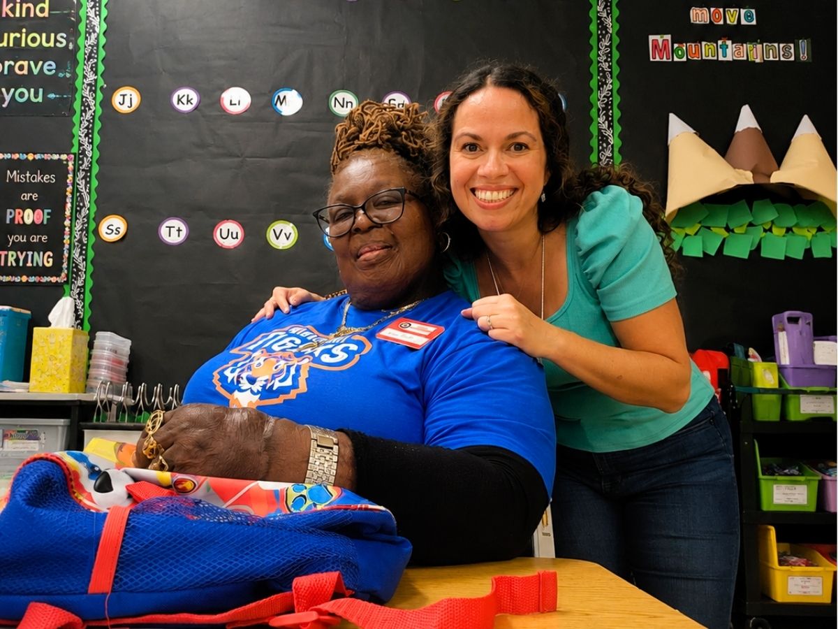 A Classroom Grandparent volunteer and elementary school teacher pose together in a classroom, highlighting the partnership that helps students build confidence and literacy skills.