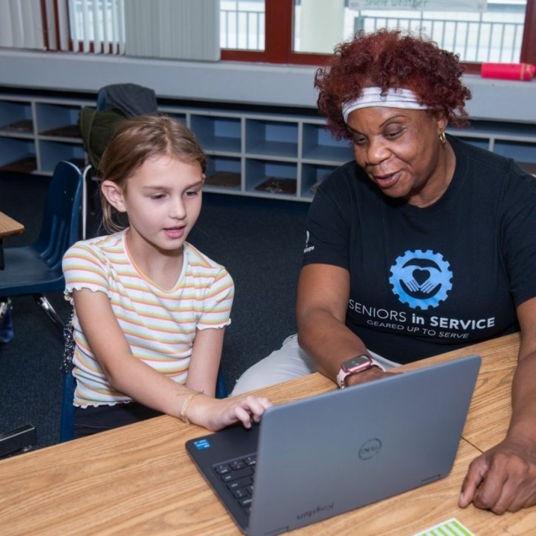 Classroom Grandparent volunteer tutors a student using a computer to practice letter recognition and phonics skills.
