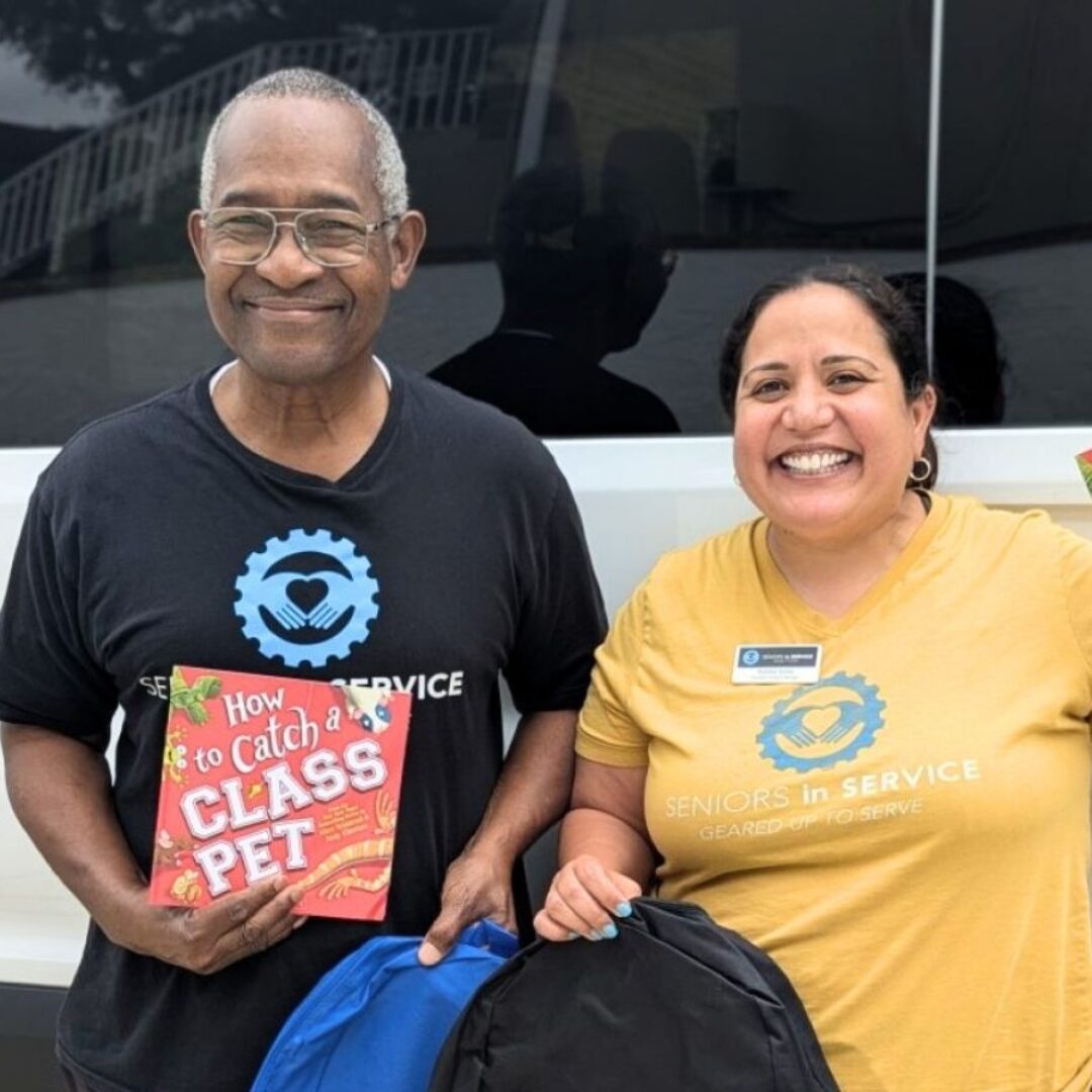 Classroom Grandparent volunteer Anthony and Seniors in Service staff member Natalie smile while holding up school backpacks and books for classrooms.