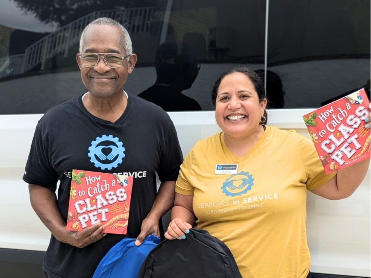 Classroom Grandparent volunteer Anthony and Seniors in Service staff member Natalie smile while holding up school backpacks and books for classrooms.