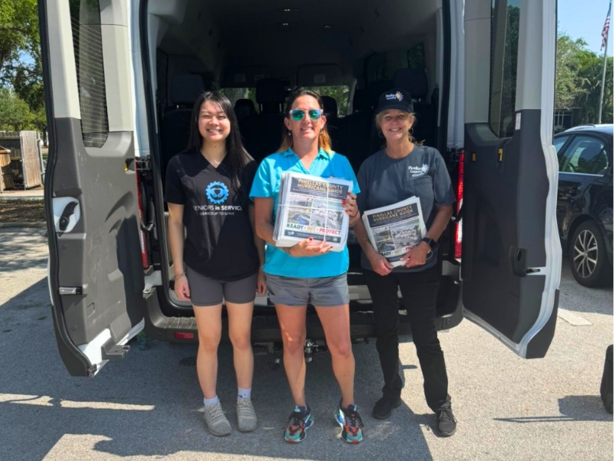 Three volunteers stand at the back of a community outreach van holding hurricane preparedness guides for older adults.