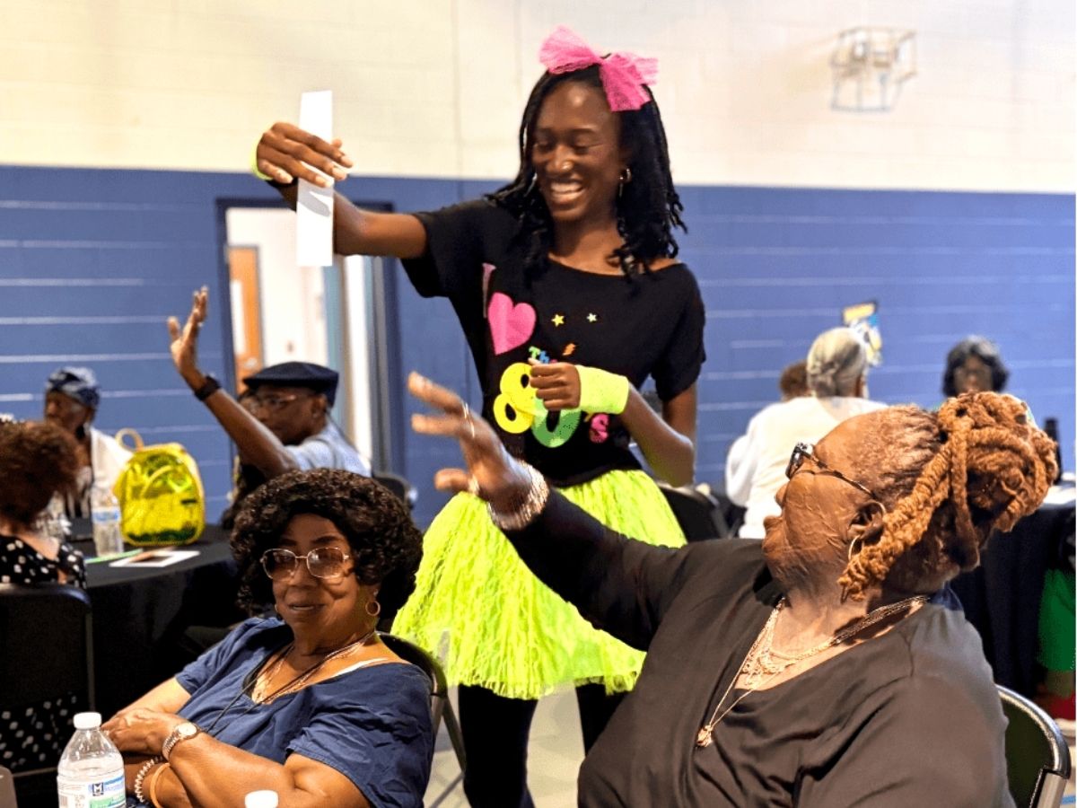 Volunteer in colorful 1980s attire hands out a game prize during Seniors in Service's 40th anniversary volunteer celebration while attendees enjoy the festivities.