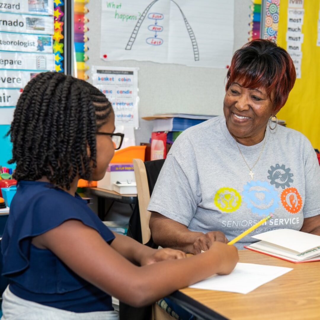 Seniors in Service Classroom Grandparent volunteer smiling with student while teaching reading comprehension.