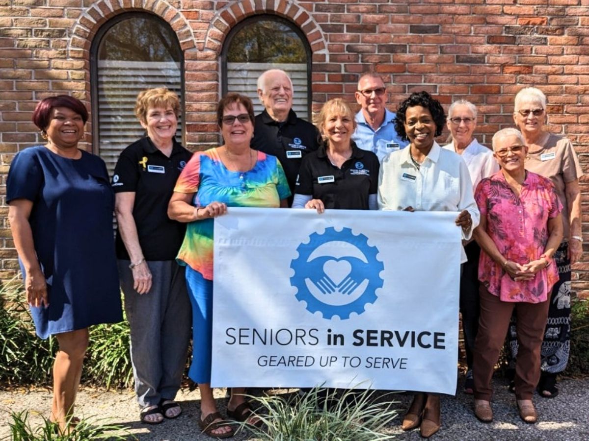 Seniors in Service Ambassadors pose together outdoors holding a Seniors in Service banner after completing ambassador certification training.