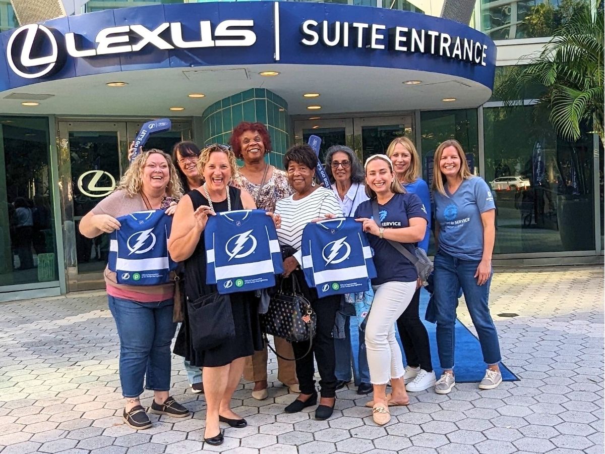 Seniors in Service staff and Classroom Grandparent volunteers smiling together outside Amalie Arena before a hockey game in Tampa Bay.
