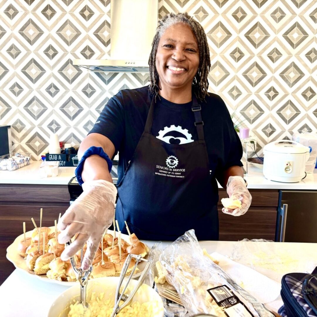 Seniors in Service volunteer smiling while preparing food for veterans during a community meal in Tampa Bay.