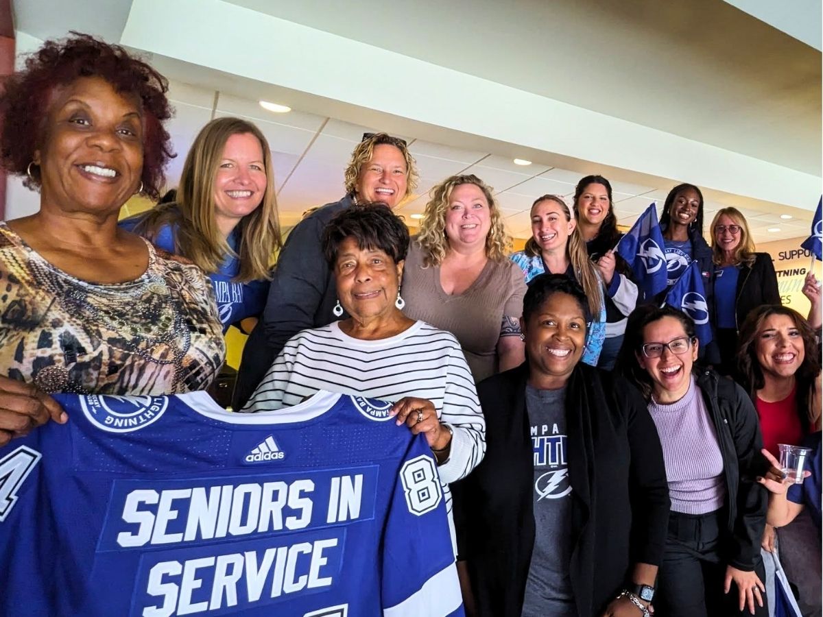 Seniors in Service staff smiling with Classroom Grandparent volunteers holding Tampa Bay Lighting merchandise at Hockey Game.
